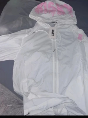 Vit vindjacka med rosa tryck - Snygg vit vindjacka från RISKY med huva och dragkedja framtill. Jackan har ett rosa tryck på huvan och ett litet rosa R på axeln. Perfekt för blåsiga dagar och streetwear-stil.