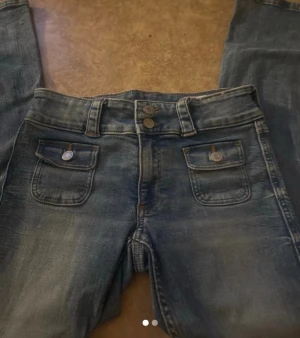 Blå jeans med ficklock och knappar - Snygga blå jeans med två framfickor med lock och dekorativa knappar. Jeansen har en klassisk femficksmodell, bälteshällor och en lätt tvättad look. Perfekta för en avslappnad stil och passar till det mesta.