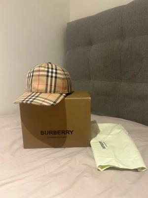 Burberry keps - Säljer en ikonisk Burberry keps i beige med det klassiska rutmönstret i svart, vitt och rött. Kepsen har böjd skärm och justerbar rem bak. Kommer med lådan och dustbag. 