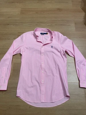 Rosa randig skjorta från Polo Ralph Lauren - Säljer en rosa och vit randig skjorta från Polo Ralph Lauren i slim fit-modell. Skjortan har långa ärmar, klassisk krage och knappar framtill. Tillverkad i bomull och har den ikoniska loggan broderad på bröstet. Den har lång passform. Köpt ny av mig, ej använd 