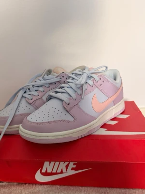 Nike Dunk Low pastell sneakers - Nike Dunk Low sneakers i pastellila och ljusblått med en ljusrosa swoosh. Skorna har klassisk låg siluett, perforerad tåbox och matchande ljusblå snören. Bara att höra av sig vid funderingar 
