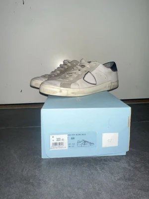 Vita Philippe Model sneakers - Snygga vita sneakers från Philippe Model med grå och svarta detaljer. Skorna har en klassisk låg silhuett, vita snören och en ikonisk sköldlogga på sidan. Tillverkade i läder och mocka för en stilren och trendig look. För fler bilder skriv till mig kan visa allt från sulan osv. Hälen har släppt lite grann och insidan missfärgning.