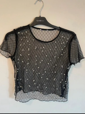 Svart mesh topp med pärlor - Säljer en svart, croppad mesh topp med korta ärmar och rund hals. Toppen har ett rutigt nätmönster och är dekorerad med vita pärlor på framsidan för en extra cool look. Perfekt att styla över ett linne eller bralette.