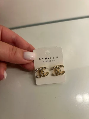 Guldiga örhängen med strass från LYN.LYN - Säljer ett par guldiga örhängen från LYN.LYN i form av två C:n som möts. Örhängena är dekorerade med gnistrande strass-stenar som ger en lyxig känsla. Perfekta för dig som gillar statement-smycken och vill sticka ut lite extra.