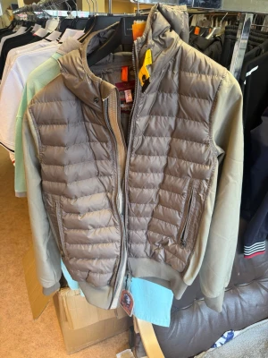 Brun hybridjacka från Parajumpers - Snygg brun hybridjacka från Parajumpers med vadderad framsida och huva. Jackan har dragkedja, två sidofickor med dragkedja och ribbade muddar. Perfekt för lager-på-lager och streetwear-stil.