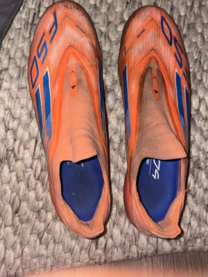 Orangea F50 fotbollsskor - Säljer ett par orangea F50 fotbollsskor med blå detaljer och klassiska ränder på sidorna. Skorna har en strömlinjeformad design utan snörning och är tillverkade i syntetmaterial för bästa känsla på planen. Perfekta för dig som vill sticka ut på matchen.