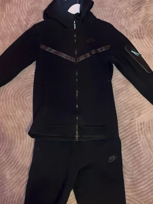 Nike Tech Fleece Set - Snyggt svart set från Nike med huva och dragkedja på tröjan samt matchande byxor. Tröjan har två fickor framtill och diskreta svarta detaljer över bröstet. Perfekt för en sportig och avslappnad stil.