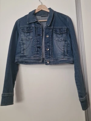 Kort blå jeansjacka från KappAhl - Snygg kort jeansjacka i klassisk blå denim från KappAhl. Jackan har långa ärmar, två bröstfickor med lock och knappar framtill. Perfekt för att lyfta din outfit med en tidlös och trendig look.