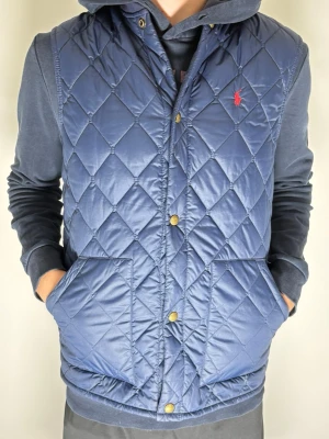 Ralph Lauren Väst Ungdom storlek XL - Tjeeena! Vi säljer nu denna unika men snygga marinblå dunväst från Polo Ralph Lauren med quiltat mönster och tryckknappar framtill. Västen har två stora fickor och en liten röd logga broderad på bröstet. Perfekt att slänga över en hoodie för en chill och stilren look. ❤️(Storleken är ungdom XL men skulle säga att den passar S/M)