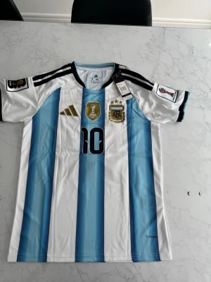 Argentina VM Messi #10 - Säljer en officiell Argentina matchtröja från Adidas med klassiska blå och vita ränder, svarta detaljer och guldiga loggor. Tröjan har nummer 10 på bröstet, FIFA World Champions 2022-märke och AFA-emblem. Tillverkad i lätt och ventilerande material.