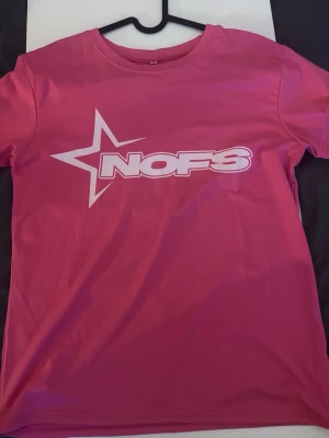 Rosa t-shirt med NOFS-tryck - Säljer en rosa t-shirt med ett stort vitt NOFS-tryck och stjärndesign på bröstet. T-shirten har rund halsringning och korta ärmar. Perfekt för dig som gillar färgstarka plagg och vill sticka ut lite extra.