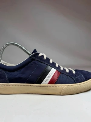 Moncler blå låga sneakers - Snygga sneakers från Moncler i marinblå mocka och textil med vita, röda och svarta detaljer på sidan. Klassisk låg modell med vita snören och beige sula. Perfekta för dig som vill ha en stilren och sportig look.