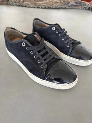 Svarta Lanvin sneakers med lackdetalj - Snygga svarta sneakers från Lanvin med ovandel i mocka och glansig lackad tå. Skorna har vita sulor och svarta skosnören. Klassisk låg modell som passar till många olika stilar. Insidan är fodrad i läder för extra komfort.