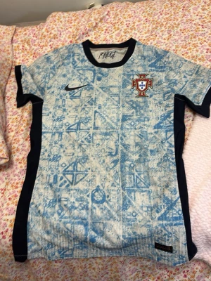 Portugal Nike fotbollströja blåmönstrad - Säljer en Portugal fotbollströja från Nike i ljusblått med unikt mönster och svarta detaljer på sidorna. Tröjan har korta ärmar, rund hals och Portugal-emblem på bröstet. Perfekt för dig som älskar fotboll och vill sticka ut på planen.