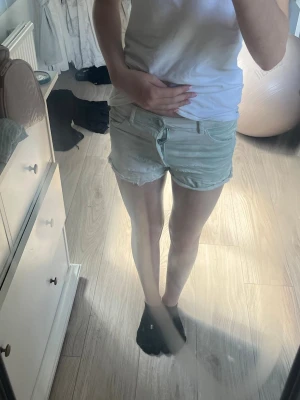 Ljusblå jeansshorts med rå kant - Säljer ett par ljusblå jeansshorts med rå, fransig kant och klassisk femficksdesign. Shortsen har knappgylf och bälteshällor, perfekt för varma dagar. Snygg och avslappnad passform som passar till det mesta.