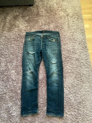 Lee Jeans - Säljer ett par blå jeans från Lee med slitningar och hål på framsidan för en cool, avslappnad look. Jeansen har klassisk femficksmodell, raka ben och tydliga slitningar på både fram- och baksida. Perfekta för dig som gillar streetstyle.
