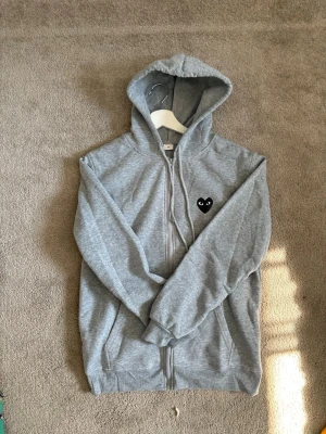 Grå Comme des Garçons PLAY-hoodie - Säljer en grå hoodie med dragkedja och huva. På bröstet finns ett svart hjärta med ögon som detalj. Hoodien har fickor framtill och ribbade muddar vid ärmar och nederkant. Perfekt för en chill och avslappnad stil.