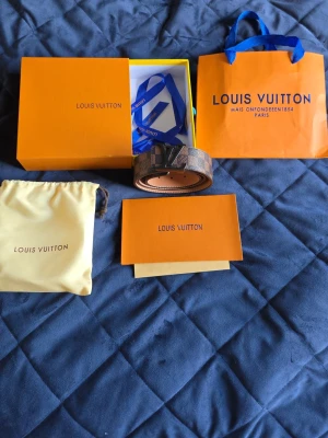 Louis Vuitton bälte rutig skärp brun - Säljer ett Louis Vuitton skärp i klassiskt rutigt mönster i brunt och svart med en stor LV-logga i metall som spänne. Skärpet kommer med originalförpackning, dustbag och påse. Perfekt accessoar för att lyfta din outfit.