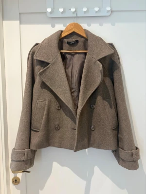 Kort brun kappa från Gina Tricot - Stilren kort kappa i brun ullmix från Gina Tricot. Dubbelknäppt med stora slag, fickor fram och dekorativa remmar vid ärmsluten. Perfekt för höst och vinter när du vill ha en trendig och varm look.