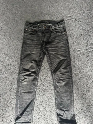 Grå slim fit jeans från Nudie Jeans - Säljer ett par grå slim fit jeans från Nudie Jeans med klassisk femficksmodell och snygga slitningar. Jeansen har en diskret tvättad look, orange sömmar och Nudie-logga på bakfickan. Perfekta för dig som gillar en avslappnad men stilren stil.