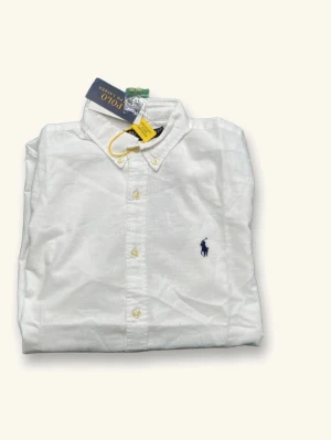Vit skjorta från Polo Ralph Lauren - Klassisk vit skjorta från Polo Ralph Lauren med knappslå och broderad logga på bröstet. Skjortan har lång ärm och är tillverkad i bomull för en skön och stilren look. Perfekt till jeans eller chinos.