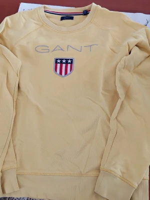 Gul sweatshirt från GANT - Gul sweatshirt från GANT med broderad logga och ett stort emblem på bröstet. Tröjan har rund halsringning, långa ärmar och ribbade muddar vid ärmslut och nederkant. Perfekt för en avslappnad och stilren look.