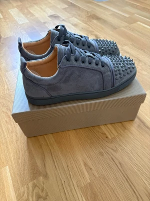 Gråa sneakers med nitar och röd sula - Säljer ett par gråa sneakers från Christian Louboutin i mocka med coola nitar på tån och den ikoniska röda sulan. Skorna har lågt skaft, svarta snören och en clean design som sticker ut. Perfekta för dig som vill ha något extra till din outfit.