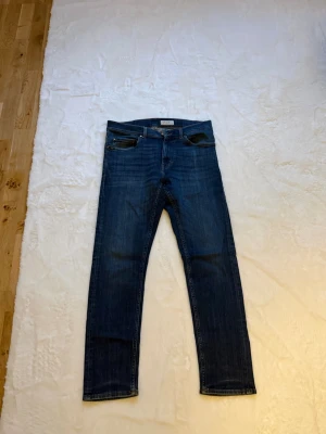 Mörkblå jeans från Tiger of Sweden - Snygga mörkblå jeans från Tiger of Sweden med klassisk femficksmodell och raka ben. Jeansen har en djup indigo färg och detaljer i guld på knappar och nitar. Perfekta för en stilren och avslappnad look.