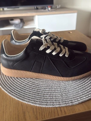 Maison Margiela gats - Svarta sneakers från Maison Margiela med beige sula och vita snören. Skorna har en mix av mocka och läder, klassisk låg modell och diskreta detaljer. Perfekta för dig som gillar stilrena och trendiga sneakers.