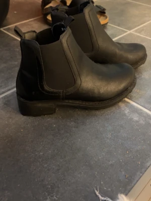 Svarta Chelsea boots i skinnimitation - Säljer ett par klassiska svarta Chelsea boots i skinnimitation med elastiska paneler på sidorna och dragflikar både fram och bak. Bootsens sula är robust och klacken är låg, vilket ger en stilren och tidlös look. Använd max 3 gånger