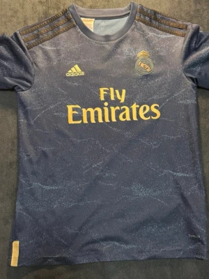 Real Madrid bortatröja Hazard #7 - Mörkblå Real Madrid bortatröja från Adidas med gul text och logga. Tryck med Fly Emirates på bröstet och Hazard #7 på ryggen. Tröjan har korta ärmar, rund hals och är tillverkad i ett lätt, ventilerande material perfekt för träning eller match.