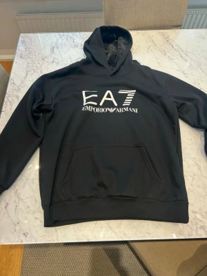 Svart hoodie från Emporio Armani EA7 - Säljer en svart hoodie från Emporio Armani EA7 med stor vit logga på bröstet. Hoodien har klassisk huva och en stor magficka framtill. Tillverkad i mjukt material som känns skönt mot huden. Perfekt för en avslappnad och trendig look.