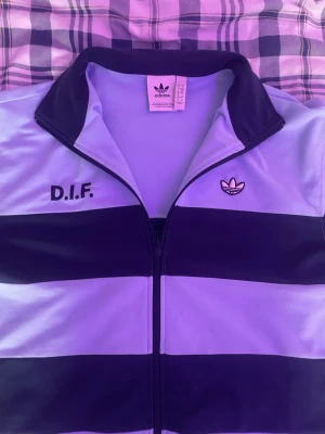 Blå adidas DIF ziptröja - Säljer en blå ziptröja från adidas med svarta breda ränder och DIF-tryck på bröstet. Tröjan har hög krage, dragkedja framtill och adidas logga broderad på ena sidan. Perfekt för dig som gillar sportig stil och Djurgården.lite stor i stoleken 