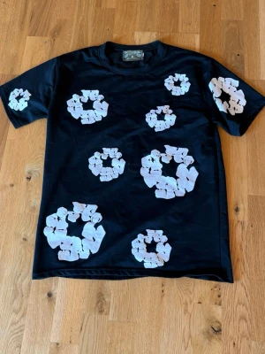 Denim tears t-shirt - Säljer en svart t-shirt från Denim Tears med stora vita blommönster tryckta över hela framsidan och ena ärmen. Klassisk passform och rund halsringning. Perfekt för dig som vill sticka ut med en unik och trendig look.