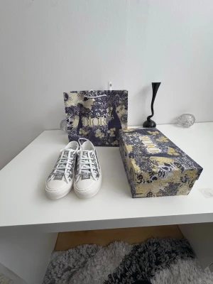 Dior gråvita sneakers med logga - Snygga sneakers från Dior i grått och vitt med tydlig Christian Dior-logga på tån och klassiskt mönster på sidan. Skorna har vita snören och sula i gummi. Perfekta för dig som vill ha en exklusiv och trendig look.