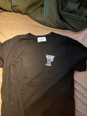 Svart Casablanca x Kaspia t-shirt - Snygg svart t-shirt från Casablanca x Kaspia med tryck på bröstet som visar en fisk och grafiska detaljer. Klassisk rund hals och korta ärmar. Perfekt för dig som gillar streetwear och vill ha något unikt i garderoben.