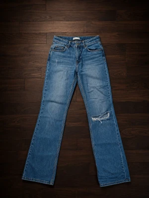 Blå bootcut jeans med slitning - Säljer ett par blå bootcut jeans med slitning på höger knä och klassisk femficksdesign. Jeansen har en avslappnad passform och är tillverkade i denim med lätt tvättad look. Perfekta för en casual och trendig stil.