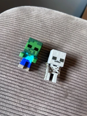 Minecraft minifigurer zombie & skelett - Två små Minecraft-figurer i plast, en grön zombie med blå byxor och en vit skelett med svart detaljering. Perfekta som samlarprylar eller dekoration till skrivbordet. Figurerna har fyrkantig form och tydliga pixlade detaljer.