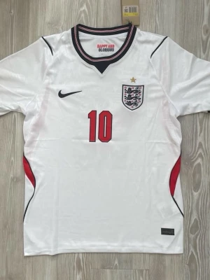 England fotbollströja Bellingham 10 - Säljer en vit England fotbollströja från Nike med röd nummer 10 och Bellingham på ryggen. Tröjan har svarta och röda detaljer, broderat landslagsemblem med stjärna på bröstet och rund hals med randig kant. Perfekt för match eller träning.