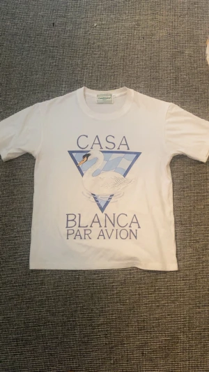Vit t-shirt från Casablanca - Vit t-shirt från Casablanca med tryck av en svan och texten 'Casa Blanca Par Avion' i blått. Klassisk rund hals och korta ärmar. Perfekt för dig som gillar stilrena plagg med unik design.
