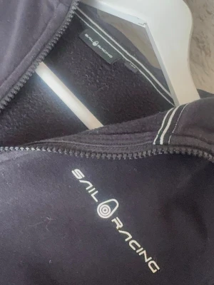 Svart hoodie från Sail Racing - Svart hoodie från Sail Racing med dragkedja framtill och vit logga på bröstet. Hoodien har huva med vita sömdetaljer och två fickor framtill. Tillverkad i mjukt material som känns skönt mot huden. Perfekt för en avslappnad och sportig stil.