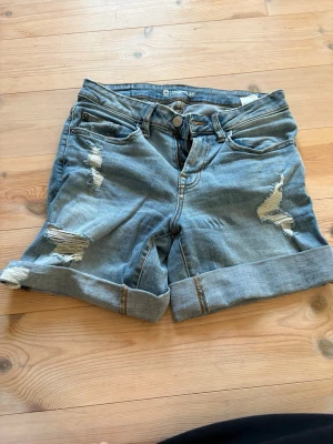 Ljusblå denimshorts med slitningar - Säljer ett par ljusblå denimshorts med uppvikta benslut och snygga slitningar framtill. Klassisk femficksmodell med knapp och dragkedja. Perfekta till sommaren för en avslappnad och trendig look.