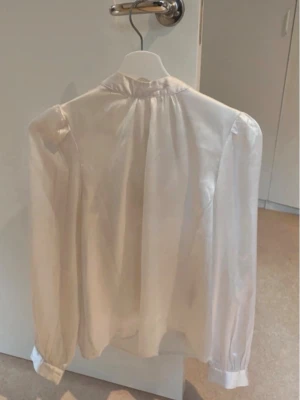 Vit transparent blus med lång ärm - Snygg vit blus i transparent material med lång ärm och klassisk krage. Blusen har en lös passform och knappar vid ärmsluten. Perfekt för en stilren och trendig look.