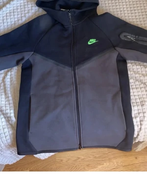 Svart Nike hoodie med dragkedja - Nike hoodie i svart och mörkgrått med hel dragkedja framtill och huva. Har en grön Nike-logga på bröstet och en praktisk ficka med dragkedja på ena ärmen. Tillverkad i mjukt och bekvämt material, perfekt för en sportig look.