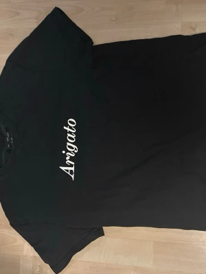 Svart Arigato t-shirt med tryck - Snygg svart t-shirt med vit Arigato-text på bröstet. Normal i storleken och bara använd ett par gånger.