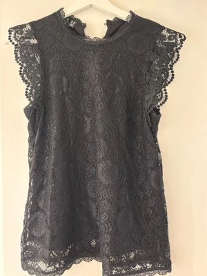 Svart spetsblus från Pieces - Snygg svart spetsblus från Pieces i storlek XS. Blusen har ett vackert spetsmönster, rund halsringning och dekorativa kanter vid ärmar och nederkant. Perfekt för dig som gillar en feminin och trendig stil.
