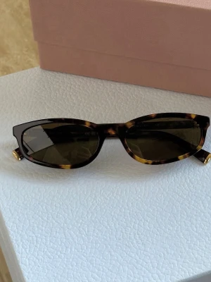 Bruna smala solglasögon från mui mui  - Snygga solglasögon med smal cat eye-form och brunmelerat sköldpaddsmönster. Glasen är mörka och bågarna har en cool retrostil. Perfekta för att lyfta din look med en trendig accessoar. Får med rosa  necessär. 