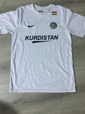 Vit Kurdistan fotbollströja Nike - Säljer en vit fotbollströja från Nike med Kurdistan-tryck på både fram- och baksidan. Tröjan har Kurdistans flagga och ett klubbmärke på bröstet. Tillverkad i lätt och ventilerande material, perfekt för träning eller match.