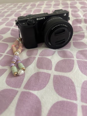 Sony A6100 systemkamera med pärlbande - Snygg svart Sony systemkamera med utbytbar lins och tydlig display på baksidan. Kameran har ett dekorativt pärlband i pastellfärger fäst på sidan. Perfekt för dig som vill ta grymma bilder och ha en personlig touch på din kamera. Helt ny skick, säljes då jag inte använt den så mycket. Aldrig tagit med mig den ut, bara testat hemma, köptes för 6 månader sedan. Skärmen har ett skärmskydd installerad och kartong och allt tillkommer. Pris kan självfallet diskuteras! Nypris: 8990