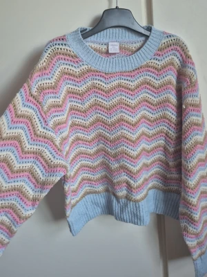 Pastellfärgad zigzag stickad tröja - Säljer en stickad tröja med zigzag-mönster i pastellfärger som ljusblått, rosa, beige och vitt. Tröjan har bred ribbstickad mudd vid hals, ärmslut och nederkant. Perfekt för dig som gillar färg. 
